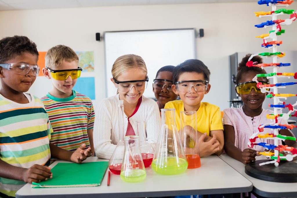 A group of kids wearing goggles are standing around the table and looking down at their lab project.