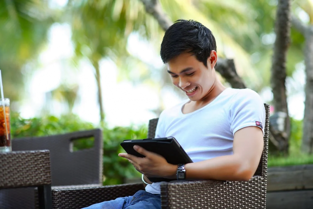 A young Asian-looking guy sits in the chair outdoors and looks into a tablet screen. 