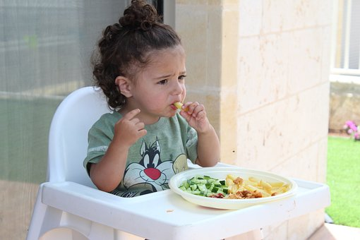 The young multi-ethnic kid sits in a baby chair, eating food off her plate. 
