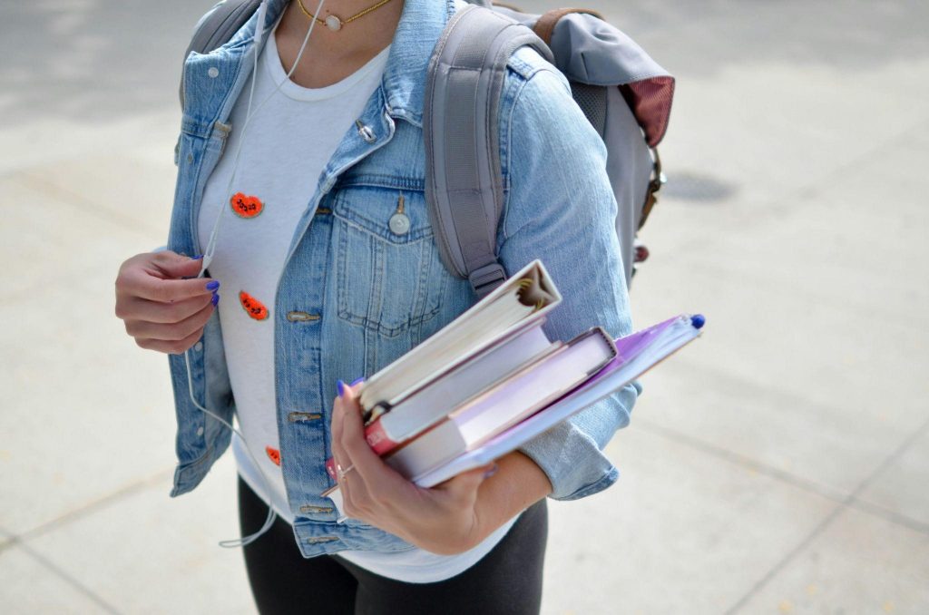 a female in a jean jacket is holding a stack of books in her left hand.