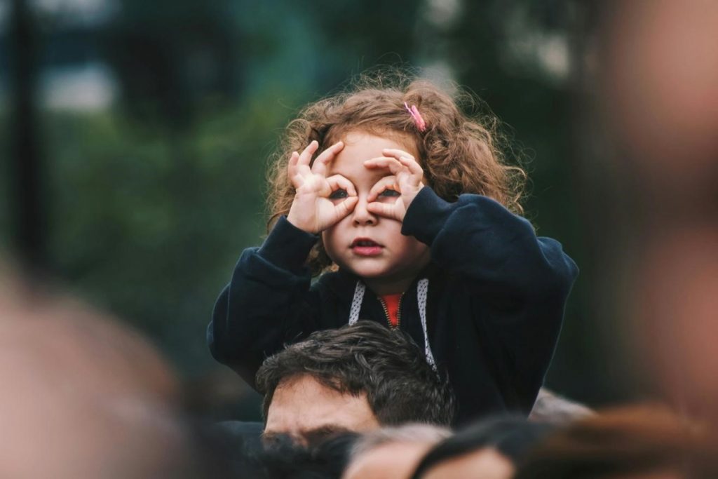 A young girl sits on her parent’s shoulders and acts as if her hands are binoculars. 