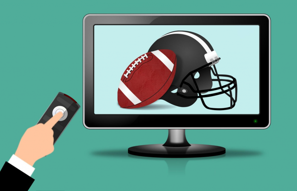 picture of a football helmet and a ball on a TV screen right next to a hand holding a remote control.