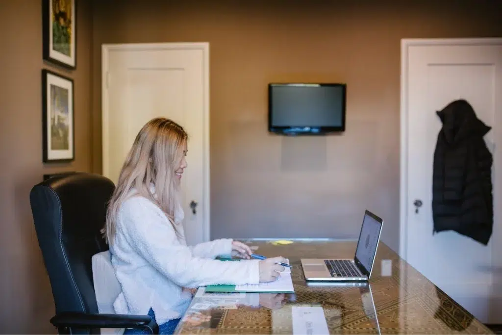 Blond-haired female sits at the table working on her laptop.