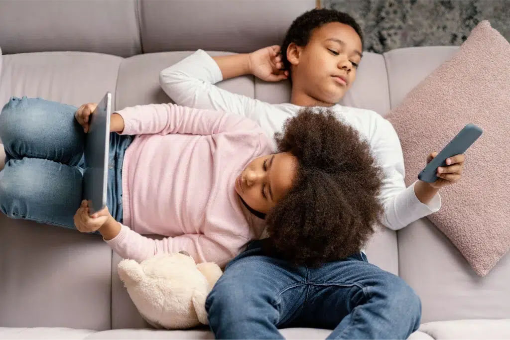 two siblings laying on the sofa entertaining themselves with gadgets