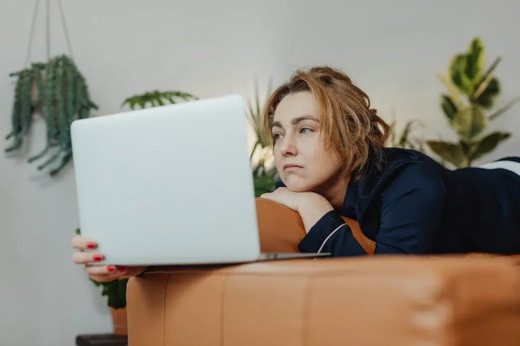 a sad looking female is laying on the sofa, looking at her computer screen