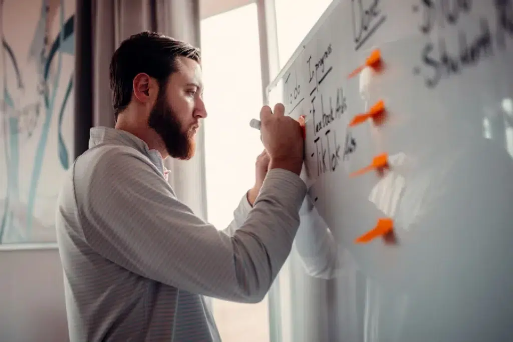 a young bearded male is writing something on a white board.