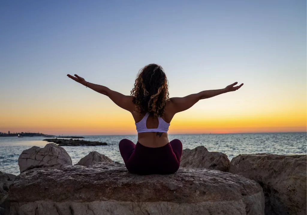 female with curly hair sits on the rocks on the beach facing the sea with her hands wide open doing yoga