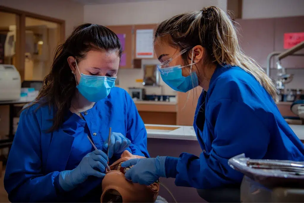 Two dental students in scrubs practice on a dummy in a classroom setting